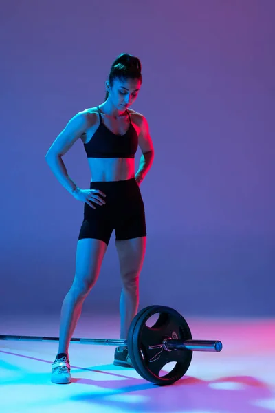Kadın sporcunun stüdyo fotoğrafı, halterli poz veren kadın neon ışıkta mor arka planda izole edilmiş. Spor, güzellik, güç kavramı — Stok fotoğraf