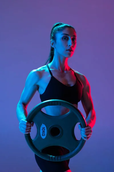 Uma mulher musculada em treinamento de sportswear com disco de barra isolado em fundo roxo em luz de néon. Esporte, beleza, conceito de força — Fotografia de Stock