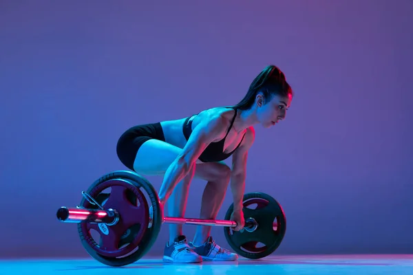 Retrato de mulher musculada em treinamento de sportswear com uma barra isolada em fundo roxo em luz de néon. Esporte, conceito de levantamento de peso — Fotografia de Stock