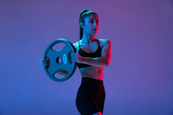 Uma mulher musculada em treinamento de sportswear com disco de barra isolado em fundo roxo em luz de néon. Esporte, beleza, conceito de força — Fotografia de Stock