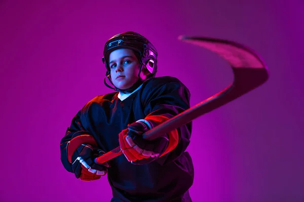 Portrait of little boy, child, hockey player in protective helmet, uniform with stick posing isolated over purple background in neon light — ストック写真