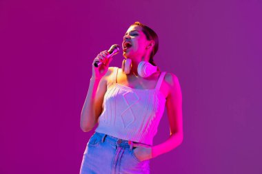 Mor arka planda, neon ışıkta, filtrede mikrofonla şarkı söyleyen güzel mutlu genç bir kızın portresi. Duyguların kavramı, müzik, yüz ifadeleri