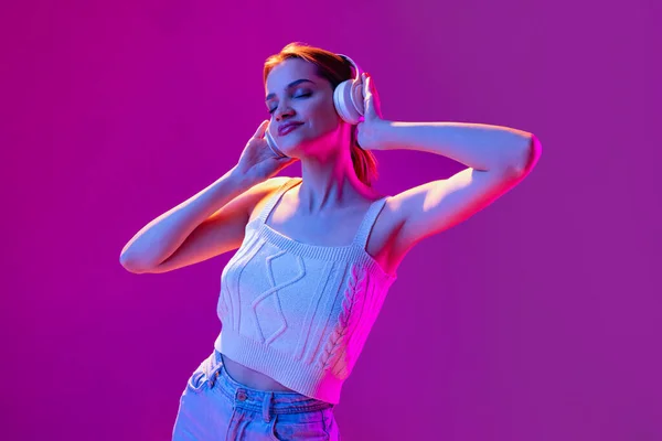 Portret młodej pięknej dziewczyny w słuchawkach słuchających muzyki odizolowanej na fioletowym tle w neonowym świetle, filtr. Pojęcie emocji, muzyki, wyrazu twarzy — Zdjęcie stockowe