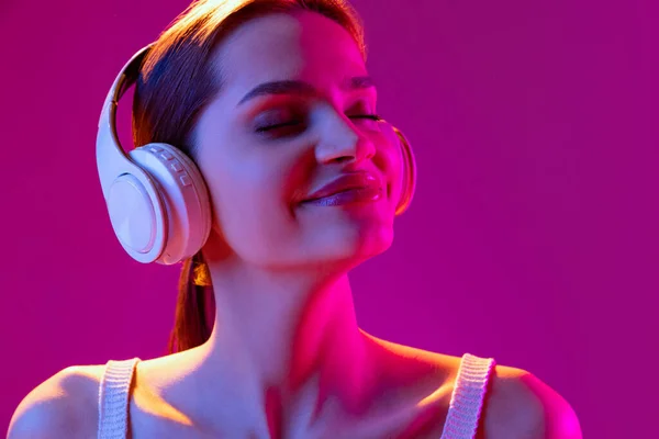 Closeup portrait of young beautiful happy girl in headphones listening to music isolated on purple background in neon light, filter. Concept of emotions, music, facial expressions — ストック写真