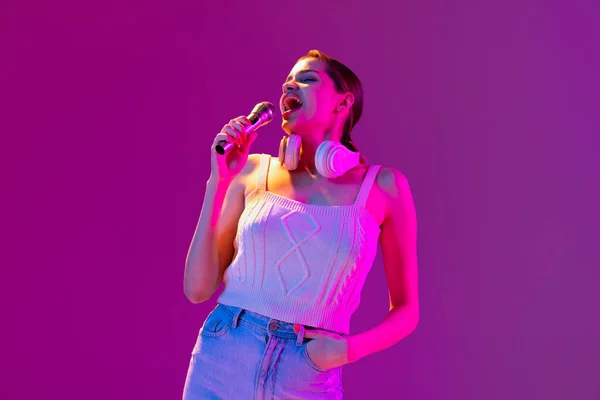 Portrait of young beautiful happy girl singing with microphone isolated on purple background in neon light, filter. Concept of emotions, music, facial expressions — Stock Photo, Image