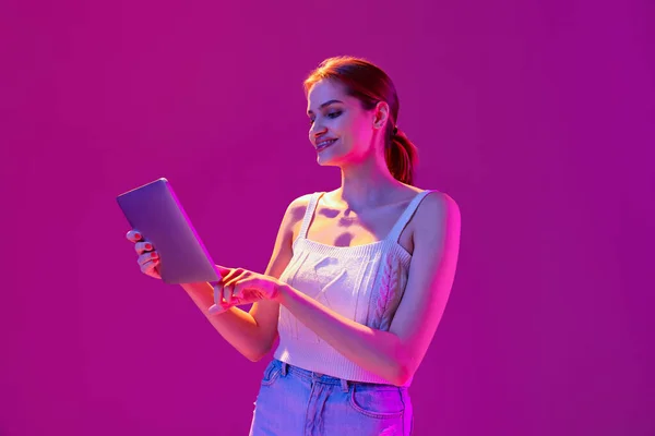 네온 필터를 사용하여 보라색 배경에 분리 된 태블릿을 사용하여 평상복을 입은 행복 한 소녀. 감정의 개념, 디지털 기술, 패션 — 스톡 사진