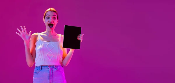 Flyer woth jovem animado menina vestindo roupas de estilo casual usando tablet isolado no fundo roxo no filtro de néon. Conceito de emoções, tecnologias digitais, moda — Fotografia de Stock