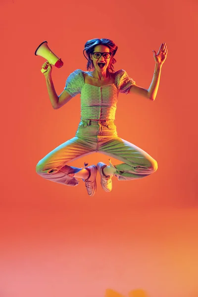 Uma menina bonita magro animado pulando com megafone isolado no fundo cor de pêssego em luz de néon, filtro. Conceito de emoções, música, notícias, informações — Fotografia de Stock