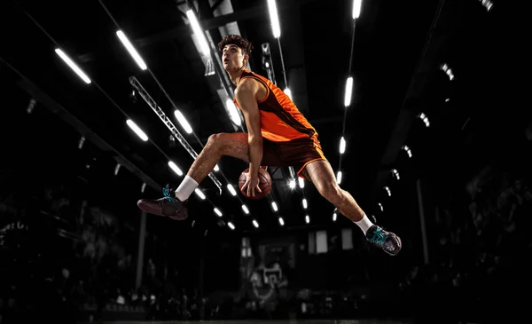 In action. Young basketball player jumping with ball in flashlights over dark gym background. Concept of sport, energy and dynamic, healthy lifestyle. Arenas drawned — 스톡 사진