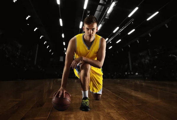 Young caucasian male basketball player posing with ball isolated over dark gym background with flashlights. Concept of motion, power, speed, healthy lifestyle, professional sport. — Foto de Stock