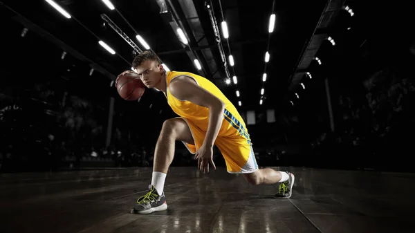 Ένας νεαρός μπασκετμπολίστας σε δράση και κίνηση σε φακούς πάνω από σκοτεινό φόντο γυμναστήριο. Έννοια του αθλητισμού, της ενέργειας και δυναμική, υγιεινό τρόπο ζωής. Αρένες τραβηγμένες — Φωτογραφία Αρχείου