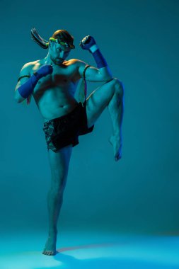 Diz tekmesi. Mavi stüdyo arka planında izole edilmiş profesyonel Tayland boksörünün dinamik portresi. Spor, Muay Thai, yarışma, dövüş kulübü konsepti