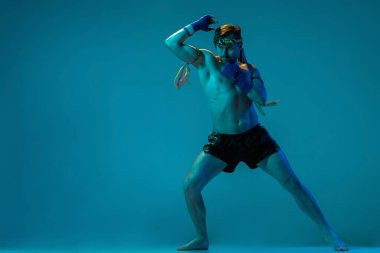 Mavi stüdyo arka planında izole edilmiş profesyonel Tayland boksörünün dinamik portresi. Spor, Muay Thai, yarışma, dövüş kulübü konsepti
