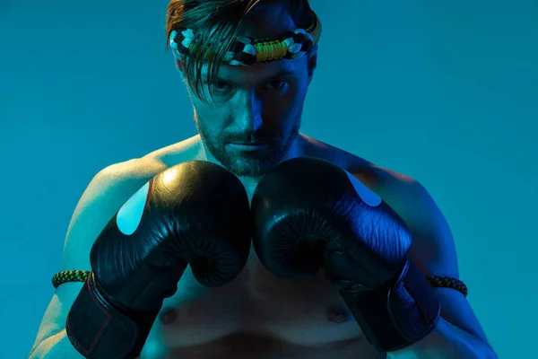 Portrait rapproché de l'homme musclé, boxeur thaï en uniforme de sport posant isolé sur fond bleu studio en néon. Sport, muay thai, compétition, concept de club de combat — Photo