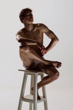 Üzücü ve ciddi görünüyor. Gri arka planda izole edilmiş yüksek sandalyede oturan beyaz şortlu, üstsüz, sportif bir genç. Sanat, moda, çeşitlilik, duygular