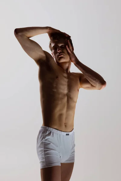 Silueta mladého svalnatého muže v bílých boxerských kalhotách, stojícího izolovaně na šedém pozadí. Přirozená krása mužského těla. Emoce, láska, péče — Stock fotografie