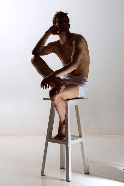 Sembra triste, serio. Un giovane uomo sportivo senza maglietta in boxer-slip bianchi seduto su seggiolone isolato su sfondo grigio. Arte, moda, diversità, emozioni — Foto Stock