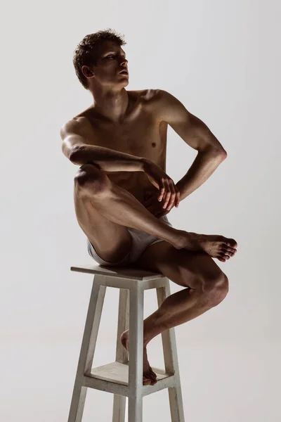Looks sad, serious. One young shirtless sportive man in white boxer-briefs sitting on high chair isolated on grey background. Art, fashion, diversity, emotions — Stock Photo, Image