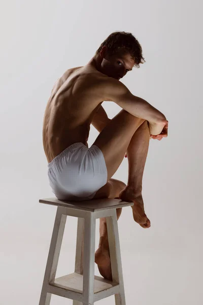 Sieht traurig aus, ernst. Ein junger, hemdloser, sportlicher Mann in weißen Boxershorts sitzt isoliert auf einem Hochstuhl vor grauem Hintergrund. Kunst, Mode, Vielfalt, Emotionen — Stockfoto