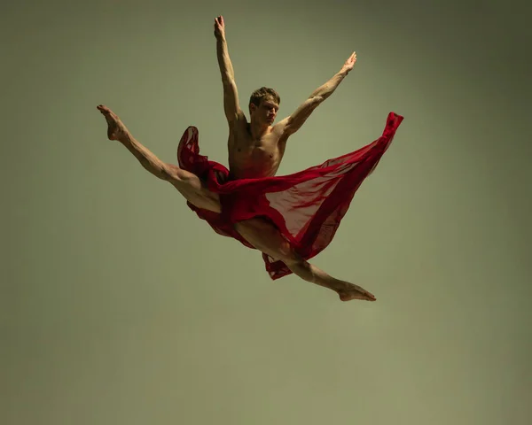 Voo. Um jovem homem musculado, dançarino de balé flexível em ação com tecido vermelho, pano isolado no fundo cor de oliva. Teatro, emoções, graça, arte, conceito de beleza. — Fotografia de Stock