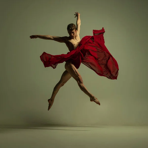 Jeden młody umięśniony mężczyzna, elastyczny tancerz baletowy w akcji z czerwoną tkaniną, tkanina izolowana na tle koloru oliwkowego. Teatr, emocje, wdzięk, sztuka, koncepcja piękna. — Zdjęcie stockowe