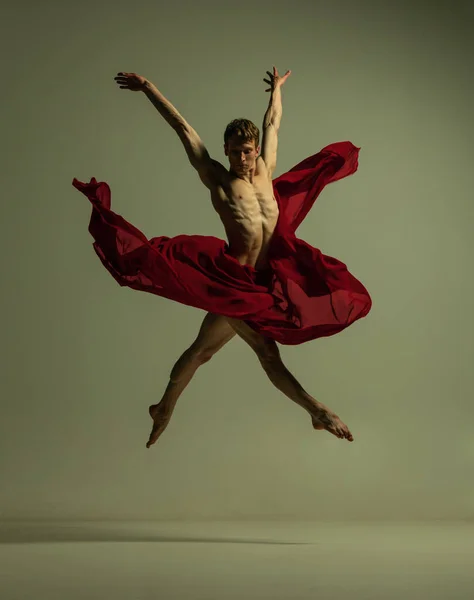 Voo. Um jovem homem musculado, dançarino de balé flexível em ação com tecido vermelho, pano isolado no fundo cor de oliva. Teatro, emoções, graça, arte, conceito de beleza. — Fotografia de Stock