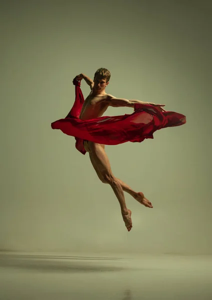 Um jovem homem musculado, dançarino de balé flexível em ação com tecido vermelho, pano isolado no fundo cor de oliva. Teatro, emoções, graça, arte, conceito de beleza. — Fotografia de Stock