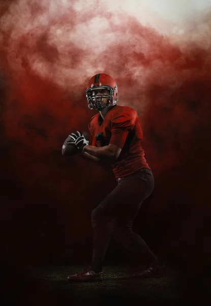 Röd himmel. Kreativt porträtt av ung man, amerikansk fotbollsspelare på stadion i rörelse på rökt bakgrund. Sport, utmaning, mål, aktivitet, idrottsliv koncept. — Stockfoto