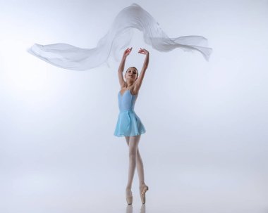 Genç bir kız, mavi elbiseli balerin olmak için can atıyor ve gri arka planda izole edilmiş kumaşla dans ediyor. Sanat, zarafet, güzellik, bale okulu konsepti