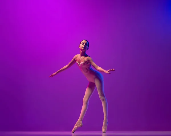 One flexible young female ballet dancer, teen in stage outfit and pointes dancing isolated on purple background in neon light. Art, grace, beauty, ballet school concept — Photo