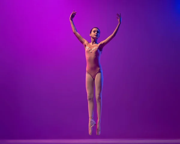 Studio shot of beautiful school age girl, ballet dancer dancing isolated on purple background in neon light. Art, grace, beauty, ballet school concept — Photo