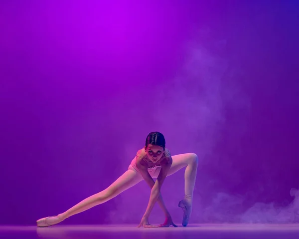 Beautiful flexible young female ballet dancer, teen in stage outfit and pointes dancing isolated on purple background in neon light with smoke. Art, grace, beauty — ストック写真