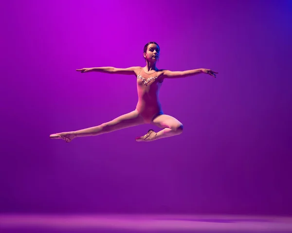 Portrait of young little ballet dancer, teen jumping isolated on purple background in neon light. Art, grace, beauty, ballet school concept — Photo