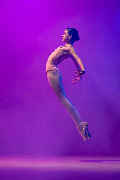 Portrait of young little ballet dancer, teen jumping isolated on purple background in neon light. Art, grace, beauty, ballet school concept — стоковое фото