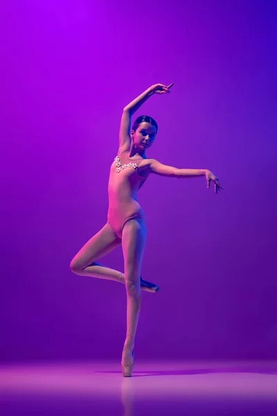 Portrait of young little ballet dancer, teen practicing, dancing isolated on purple background in neon light. Art, grace, beauty, ballet school concept — стоковое фото