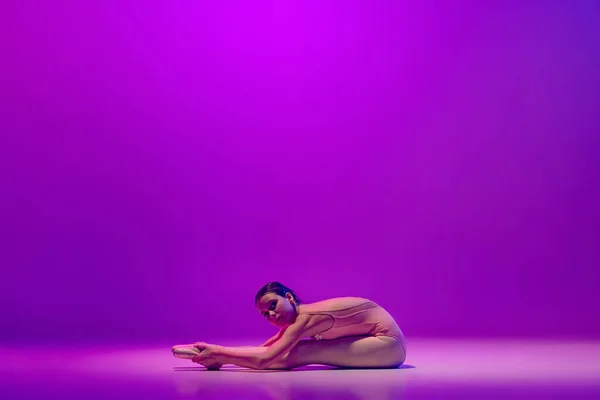 一位穿着舞台服的年轻而灵活的女芭蕾舞蹈家，坐在地板上，在霓虹灯下与紫色背景隔离。艺术、优雅、美、芭蕾舞学校概念 — 图库照片