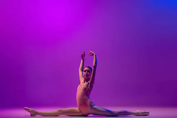 One flexible young female ballet dancer, teen in stage outfit and pointes dancing isolated on purple background in neon light. Art, grace, beauty, ballet school concept — Photo