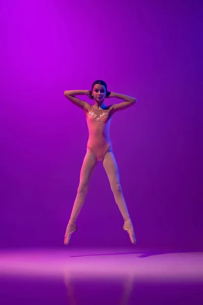 Portrait of young little ballet dancer, teen practicing, dancing isolated on purple background in neon light. Art, grace, beauty, ballet school concept — Photo