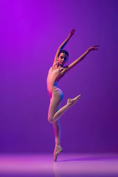 Studio shot of beautiful school age girl, ballet dancer dancing isolated on purple background in neon light. Art, grace, beauty, ballet school concept — стоковое фото