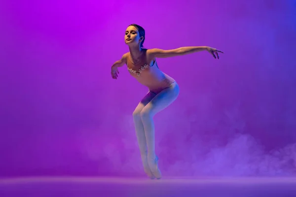 Beautiful flexible young female ballet dancer, teen in stage outfit and pointes dancing isolated on purple background in neon light with smoke. Art, grace, beauty — Photo