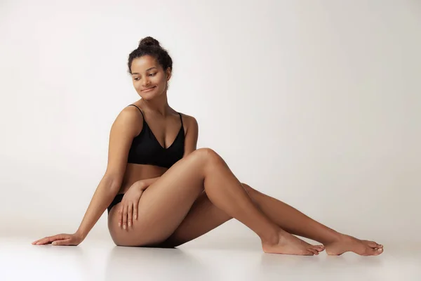 Portrait of young beautiful woman sitting on floor, posing in black underwear isolated over gray backgoround. Anti-cellulite care — ストック写真