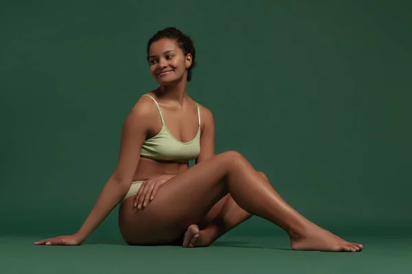 Портрет молодой нежной женщины, сидящей на полу, позируя в нижнем белье изолированы на зеленом фоне. Тело-позитивность — стоковое фото