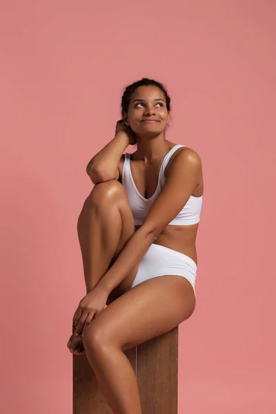 Portrait of young beautiful slim woman sitting, posing in white cotton underwear isolated over pink studio background. Bosy positivity concept — стоковое фото