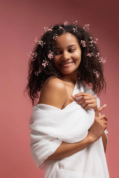 Portrait of young tender woman with small flowers in hair posing in white bathrobe isolated over pink studio background — стоковое фото