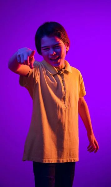 Portrét emotivního dítěte smějícího se, posměšného, ukazujícího prstem na purpurové pozadí v neonovém světle — Stock fotografie