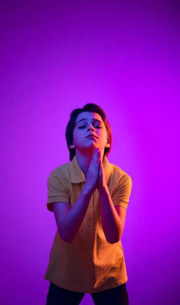 Ritratto di ragazzo emotivo, bambino che ha le mani in posizione di preghiera, posa isolato su sfondo viola in luce al neon — Foto Stock