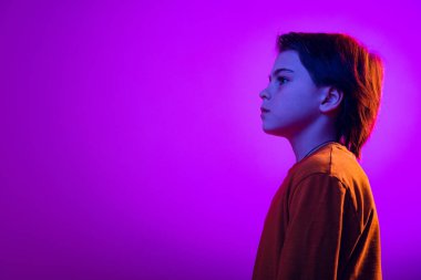 Stüdyo portresi, sakin bir ifadeyle başka yere bakan çocuk, mor arkaplanda, neon ışıkta izole edilmiş bir poz.