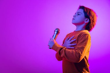 Ciddi görünümlü bir çocuğun portresi, mikrofonlu bir çocuk, elini kalbimin üzerine koyuyor, pembe arka planda, neon ışıkta izole edilmiş.