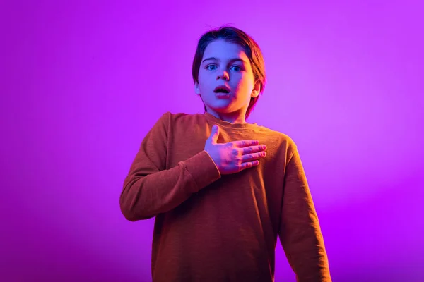 Portrait of boy, child holding hand on chest, showing emotion of fear isolated over purple studio background in neon — Foto de Stock