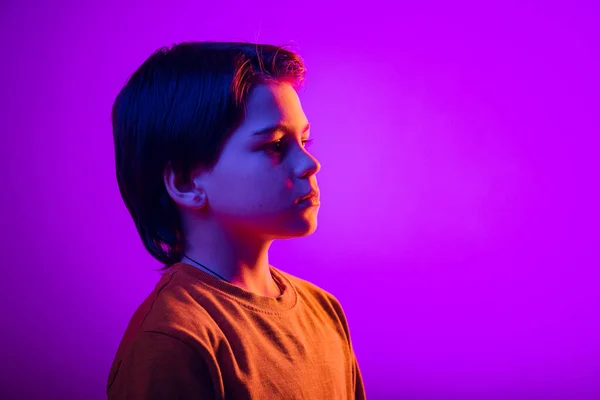 Close-up studio portrait of boy, child looking away with serious calm expression, posing isolated over purple background in neon — Foto de Stock